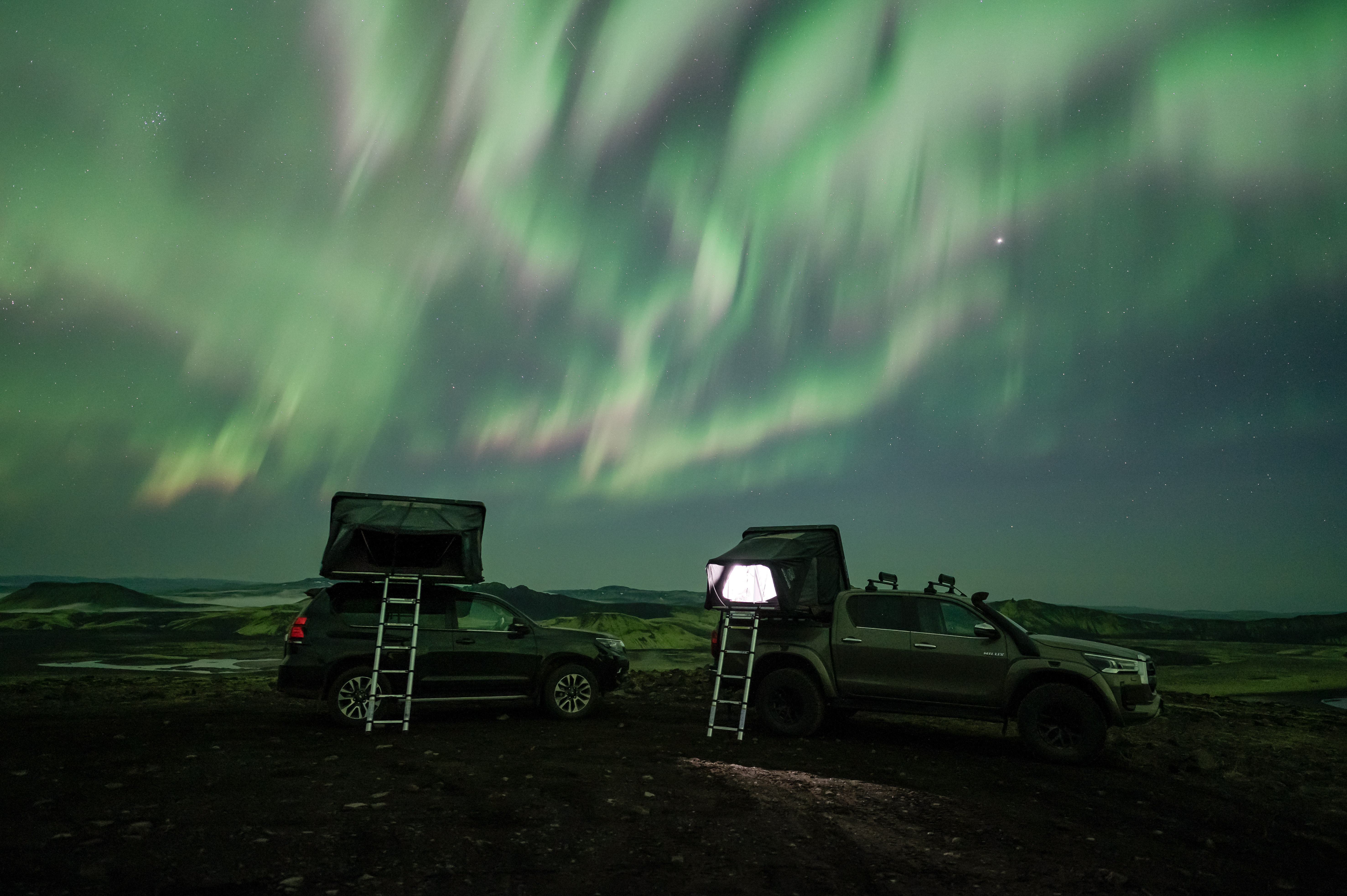 The Northern Lights dancing above two Toyota jeeps with rooftop tents in Iceland.